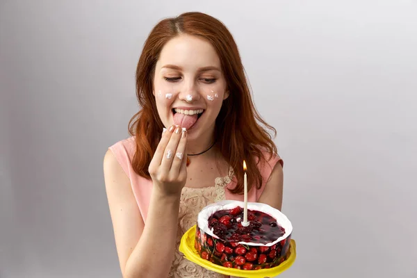 Młode piękne redhead dziewczyna niechlujstwa zjada podarował jej tort ze świecą w jej ręce, Lizanie krem z jej rąk — Zdjęcie stockowe
