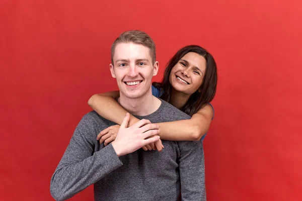 Feliz hermosa pareja sonriendo ampliamente a la cámara que muestra los dientes blancos. Amantes abrazándose en el fondo rojo aislado — Foto de Stock