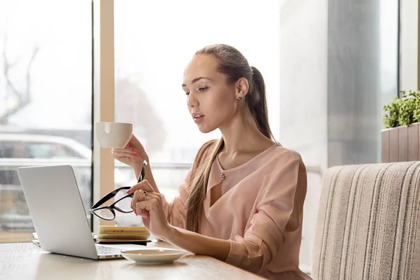 Λεπτός μοντέρνος νέος ελκυστικού επιχειρηματικού κυρία blogger με μακριά μαλλιά που κάθεται σε ένα τραπέζι σε ένα καφέ με φορητό υπολογιστή και το φλιτζάνι του καφέ — Φωτογραφία Αρχείου