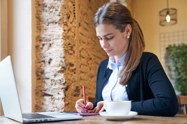 Ritratto di donna adulta che lavora nel loro business a un tavolo in un bar prendendo appunti in un taccuino sopra una tazza di caffè e laptop — Foto Stock