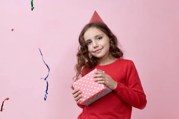 Alegre niña feliz sostiene caja con regalo. Cabello largo y ondulado, suéter rojo y sombrero de fiesta en su cabeza. Retrato sobre fondo rosa . —  Fotos de Stock