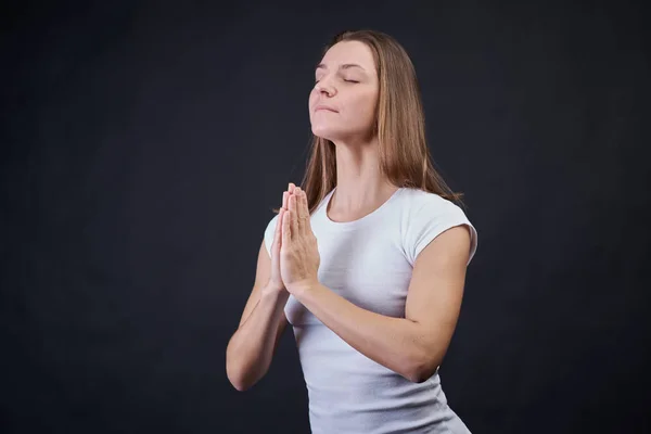 Ordinary blonde woman in white plain t-shirt on isolated black background holding hands in prayer and closed eyes. The girl of European nationality concentrates and learns emotional balance. Royalty Free Stock Photos