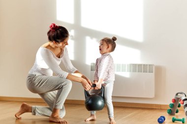 küçük şirin kız kaldırmak ağır ağırlık çalışmakla iş ve eğlenceli ifade ile. Annesi komik resim gülümsüyor ve yardımcı olur