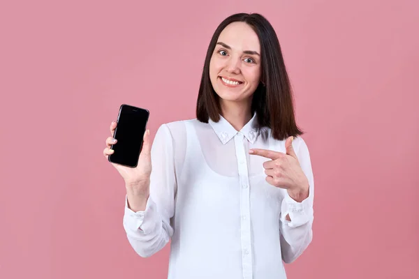 Ung trevlig brunett flicka vänligt ler brett, pekande fingret på smartphone i handen. — Stockfoto