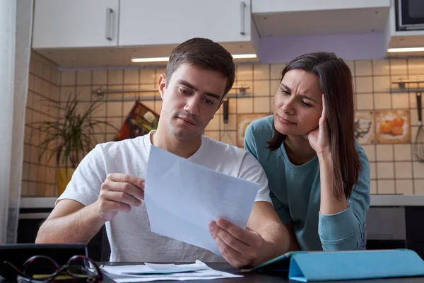 Молодая пара, сидящая за столом на кухне, понимает финансы молодой семьи, изучает бухгалтерские и налоговые документы — стоковое фото