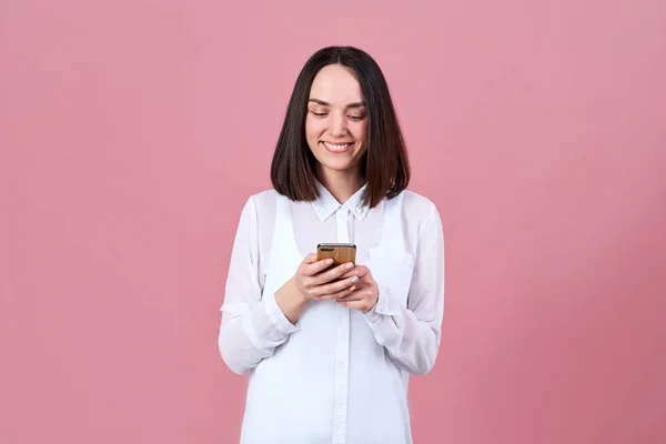 En ung attraktiv vänliga brunett kvinna i en vit skjorta ler brett med tänderna och skriver ett meddelande på sin smartphone. — Stockfoto
