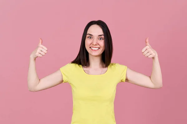 Positiv vänlig brunett tjej ler brett och visar fingrarna upp. Studio porträtt av en kvinna i gul t-shirt — Stockfoto