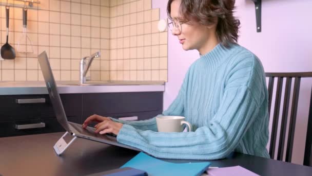 Vídeo da menina olha monitor de tela, digitando texto no teclado, escrevendo atentamente, trabalhando, sentado em quarentena durante a pandemia. Linda morena Bob cabelo usa camisola azul. caneca de café Bloco de notas — Vídeo de Stock