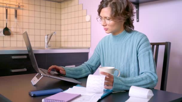 若い女性のビデオ,請求書を見て,家賃学校のためのすべての債務を支払う方法について考えて.ボブの髪型と美しいブルネットの女の子は青いセーターを着ています。テーブルの上にノートパソコン、コーヒーマグカップメモ帳ホーム — ストック動画