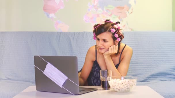 Hermosa chica está viendo una emocionante serie de televisión beber refrescos y sentarse en el sofá. Escribir texto trabajando en el ordenador portátil. Días divertidos durante la cuarentena. mujer lleva parches chemise bajo los ojos rulos de pelo — Vídeos de Stock