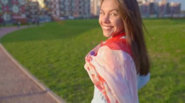 Kadın döndü, profili, nazik gülümsemesi, kameraya baktı, yarım yamalak, güzel gözlü. Güneşli bir videoda kadın gün batımında parkta yürürken görülüyor. Orta yaşlı esmer kız, rüzgar uzun siyah saçlar savuruyor.