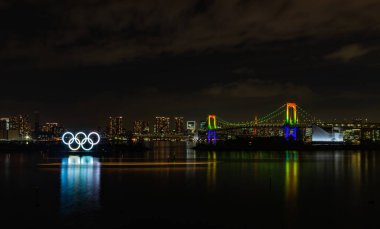 Tokyo Körfezi 'nin bir resmi, aydınlatılmış Gökkuşağı Köprüsü, ve aydınlatılmış Olimpiyat Halkaları, geceleri.