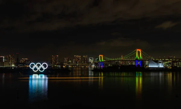 Tokyo Körfezi 'nin bir resmi, aydınlatılmış Gökkuşağı Köprüsü, ve aydınlatılmış Olimpiyat Halkaları, geceleri.