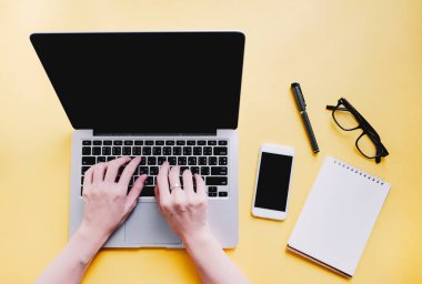 Çalışma masasında kadın eli bilgisayarda akıllı telefon, kağıt ve gözlükleri parlak sarı arka planda, iş ve yaşam tarzı kavramında daktilo eden düz bir masa.