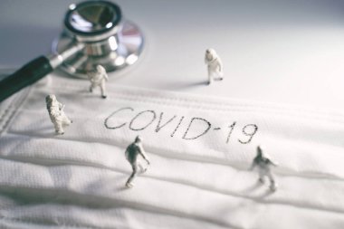 Koruyucu kıyafetli minyatür doktorlar Covid-19 ve Coronavirus salgınlarını önlüyor. Üzerinde stetoskopla yazılmış Covid-19 yazılı cerrahi maske.