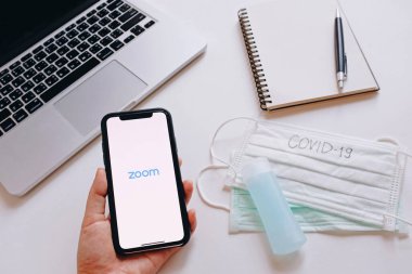 BANGKOK, THAILAND - 28 Mart 2020: Çalışma masasının düz yatağı ve kadın akıllı telefonu yakınlaştırma uygulaması ile birlikte metni covid-19 metni ile maskelerin yanında tutuyor.