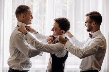 stylish groomsmen helping happy groom getting ready in the morning for wedding ceremony. luxury man in suit in room. clipart