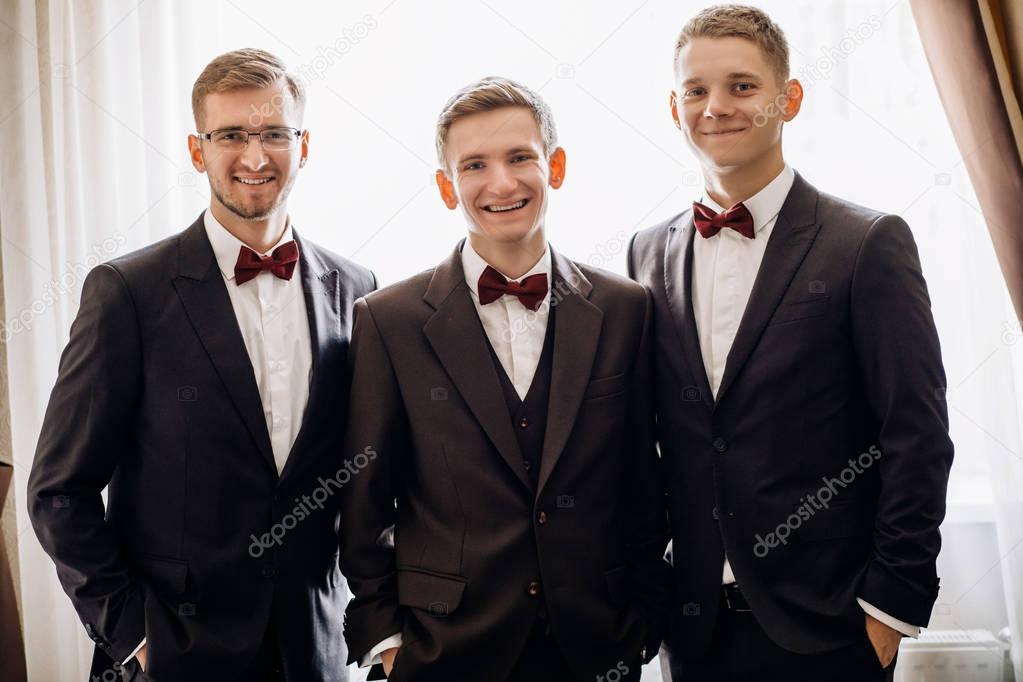 stylish groom with groomsmen in suits with boutonniere posing, getting ready in morning for wedding ceremony
