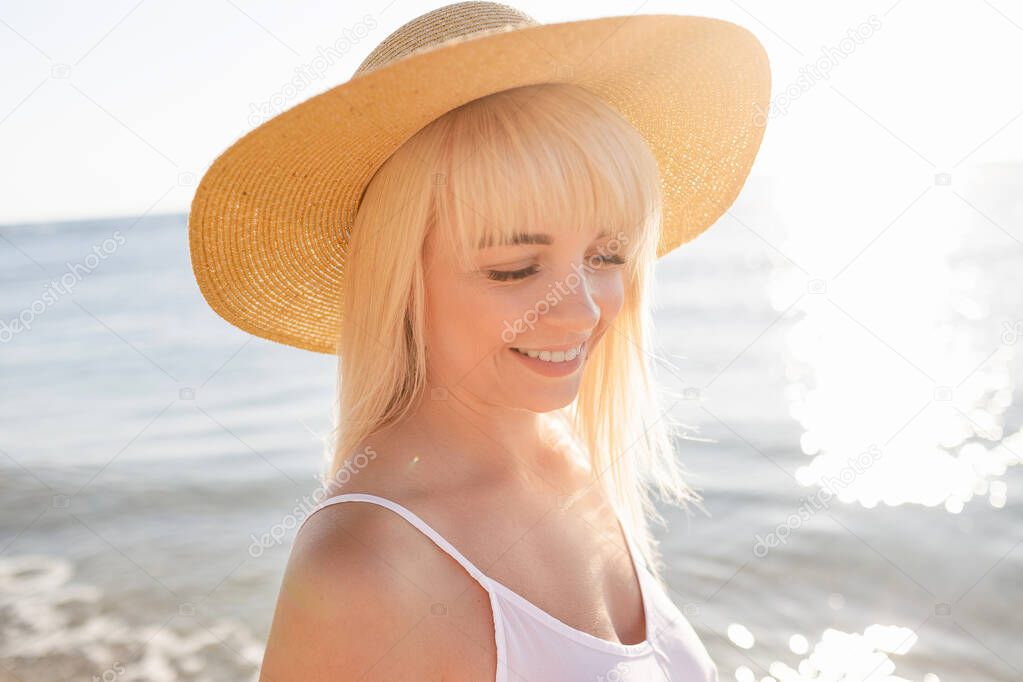 Beautiful blonde woman in white dress and straw hat on the beach
