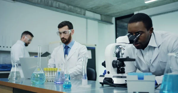 Δύο Πολυεθνικοί Επιστήμονες Εργαστηρίου Μιλάνε Ενώ Ένας Δουλεύει Στο Μικροσκόπιο — Φωτογραφία Αρχείου
