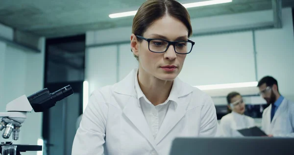 Close up of the beautiful young Caucasian woman in glasses working in the laboratory and looking in the microscope, then typing on the laptop computer. Camera zooming out.