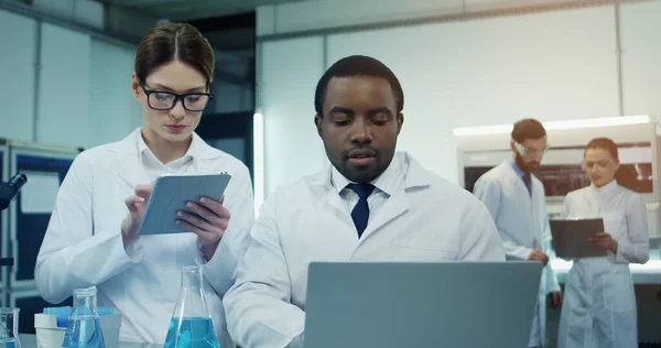African American man and Caucasian woman working and talking in the laboratory while he sitting at the laptop and she standing with a tablet computer next to him.
