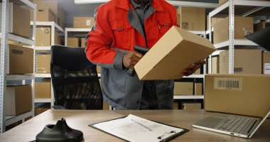 Postanedeki dizüstü bilgisayarda paketlerle çalışan üniformalı Afro-Amerikalı genç adam. Postacı tarayıcı ile karton kutuyu tarıyor