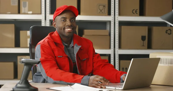 Młody, wesoły afroamerykański listonosz w czerwonej iniformie i czapce siedzi przy biurku w sklepie pocztowym i pracuje przy komputerze. — Zdjęcie stockowe