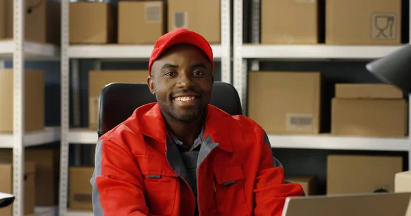 Νεαρός χαρούμενος Αφρο-Αμερικανός ταχυδρόμος σε κόκκινο iniform και καπάκι κάθεται στο γραφείο στο κατάστημα ταχυδρομικών γραφείων και εργάζεται στον υπολογιστή. — Φωτογραφία Αρχείου