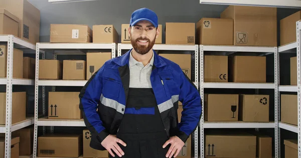 Branco jovem alegre homem de uniforme e boné sorrindo alegremente para a câmera com caixa de papelão na loja de correios . — Fotografia de Stock