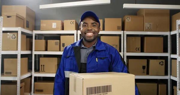 Afro-americano jovem carteiro alegre em uniforme e boné sorrindo alegremente para câmera com caixa de papelão na loja de correios . — Fotografia de Stock