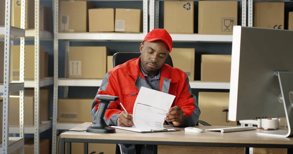 Carteiro afro-americano fardado a trabalhar num computador nos correios com encomendas. Carteiro registando caixa de correio — Fotografia de Stock