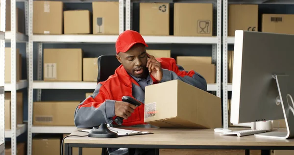 Homem afro-americano falando no celular enquanto trabalhava na loja de correios e no computador. Código de barras de digitalização do carteiro com scanner, caixa de registro — Fotografia de Stock