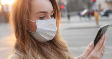 Aşağıdan, açık havada akıllı telefonunu kullanarak tıbbi maske takan güzel, beyaz bir kadın görünüyor. Caddedeki salgın sırasında telefonda mesaj atan ve haber sayfasını kaydıran güzel kız..