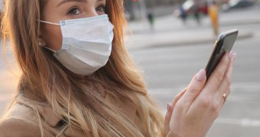 Tıbbi maske takan, mesaj atan ve caddede cep telefonunu dinleyen beyaz bir kadın. Açık havada akıllı telefondan SMS yazan ve onunla sohbet eden bir kız görüyorum. Virüs konsepti