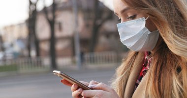 Tıbbi maskeli beyaz bir kadın akıllı telefondan web kamerasıyla konuşuyor ve parmağıyla el sallayarak hareket etmiyor. Evde kal ve dışarı çıkma konsepti yapma. Salgın konsepti