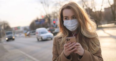 Tıbbi maskeli beyaz bir kadın yolda ve arabalarla sokakta mesaj atıyor ve cep telefonlarını dinliyor. Açık havada akıllı telefondan yazı yazan ve kaydıran bir kız. Salgın sağlık konsepti.