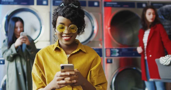 Chica afroamericana bebiendo jugo de naranja con paja, mensaje de texto en el teléfono y esperando a que la ropa se limpie Mujer bebiendo bebida en la sala de servicio de lavandería — Foto de Stock