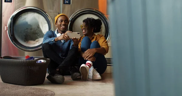 Afro-americano jovem casal sentado no chão com cesta de roupas sujas e assistir vídeo no smartphone enquanto máquinas de lavar roupa trabalhando — Fotografia de Stock