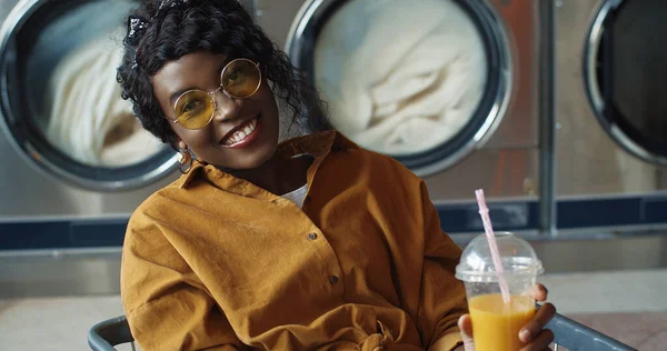 Όμορφη και χαρούμενη Αφρο-Αμερικανίδα που κάθεται σε τρόλεϊ και πίνει χυμό πορτοκαλιού με καλαμάκι, ξεκουράζεται και περιμένει να πλυθούν τα ρούχα της. — Φωτογραφία Αρχείου