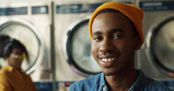 Primer plano de joven afroamericano guapo en sombrero amarillo sonriendo alegremente a la cámara en la sala de servicio de lavandería. Retrato de chico feliz riendo con lavadoras en el fondo . — Foto de Stock