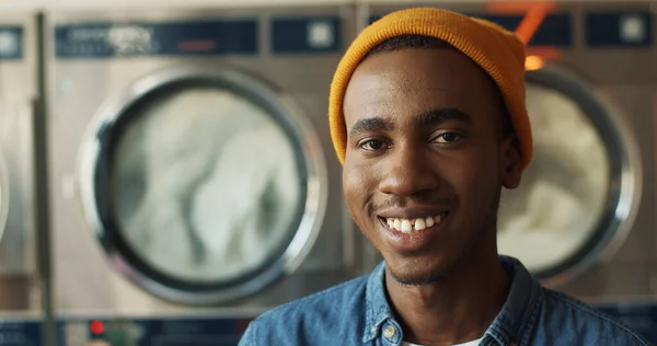 Primer plano de joven afroamericano guapo en sombrero amarillo sonriendo alegremente a la cámara en la sala de servicio de lavandería. Retrato de chico feliz riendo con lavadoras en el fondo . — Foto de Stock