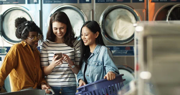 Caucasian girl showing photos on smartphone to mixed-races female friends while washing machines working and cleaning clothes. Multiethnic women watching video on phone in laundry service. — Stock Photo, Image