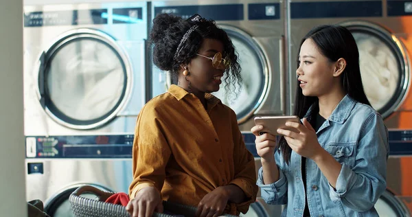 Többnemzetiségű, stílusos fiatal lányok beszélnek és néznek fényképeket vagy videókat okostelefonon. Barátok állnak a mosodában. Afrikai-amerikai és ázsiai nők telefonnal, miközben mosógépek dolgoznak. — Stock Fotó