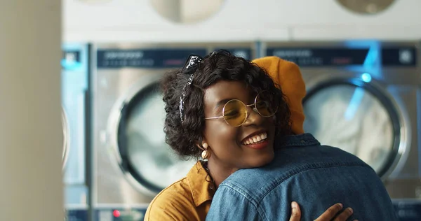 Detailní záběr Afroameričanky mladé dívky a chlap setkání a objímání v prádelně. Pár přátel setkat, mluvit a usmívat se v umývárně, zatímco pračky pracují. — Stock fotografie