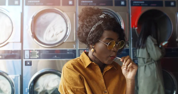 Afrikai amerikai fiatal gyönyörű lány sárga szemüvegben ül a mosodában, és lapozgatás oldalak napló. — Stock Fotó