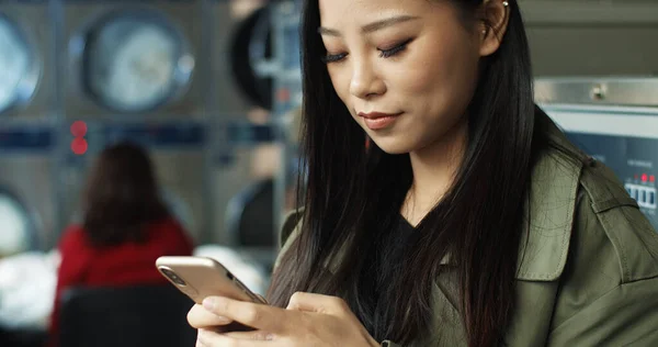 Primer plano de mujer bonita asiática con el pelo largo oscuro golpeando y mensaje de texto en el teléfono inteligente, mientras que de pie en la sala de servicio de lavandería . —  Fotos de Stock
