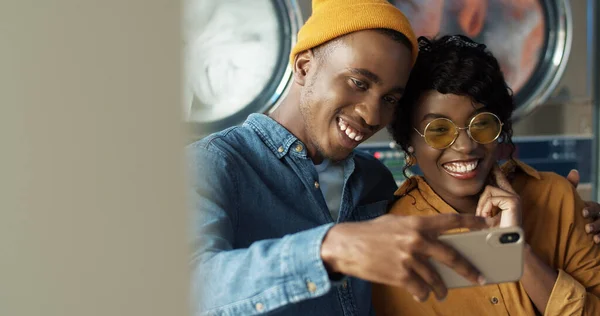 Šťastný africký americký pár v lásce objímání a úsměv na smartphone fotoaparát při pořízení selfie fotografie v prádelně. — Stock fotografie