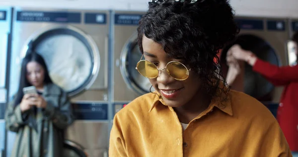 African American young pretty and stylish girl in yellow glasses standing in laundry service room and tapping on smartphone. — Stock Photo, Image