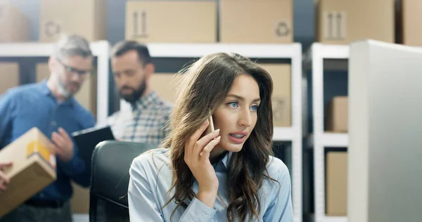 Beautiful young woman post office worker talking on mobile phone while working at computer. Female speaking on smartphone.